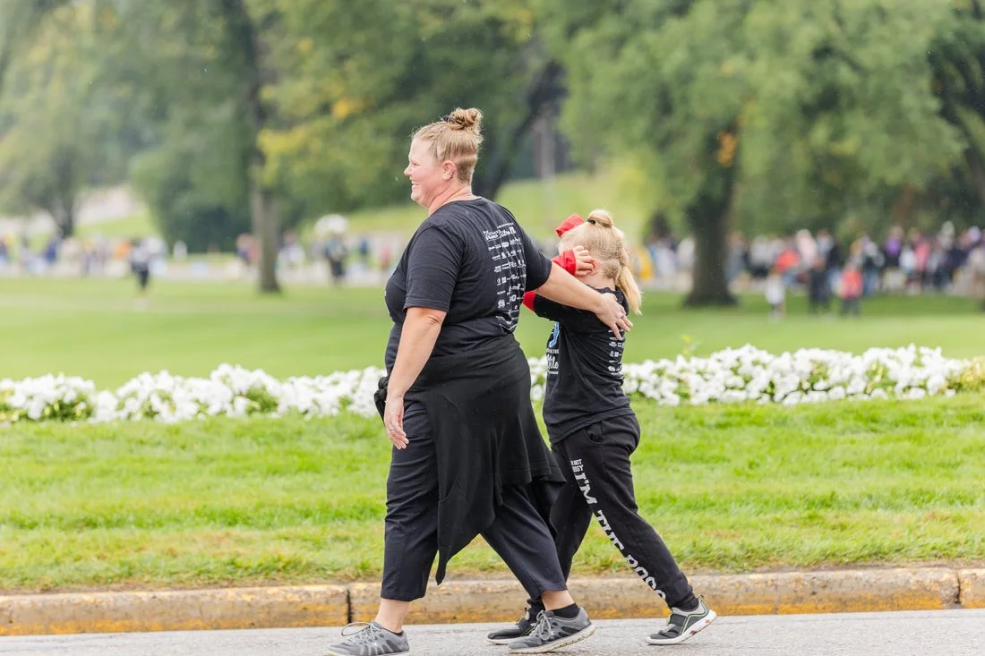 A woman and a girl, both dressed in black, are walking outdoors during daytime, smiling and hugging each other, on a grassy area with trees and flowers in the background.