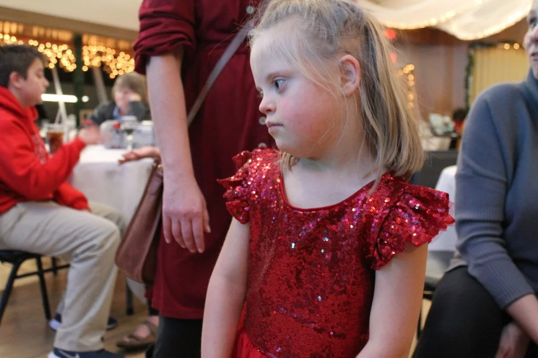 A young girl with blonde hair in a red sequin dress looking to her side at a party or celebration, with other children and adults seated at tables in the background.