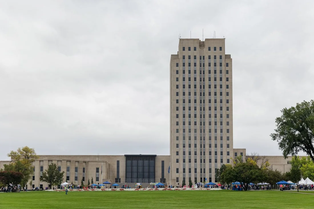 A tall beige office building with multiple windows, surrounded by a green lawn and some trees, with tents and people visible at the base on a cloudy day.