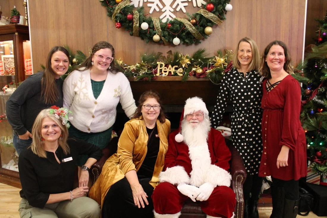 Six women and Santa Claus sitting and standing in front of a Christmas fireplace and tree, smiling at a holiday gathering.