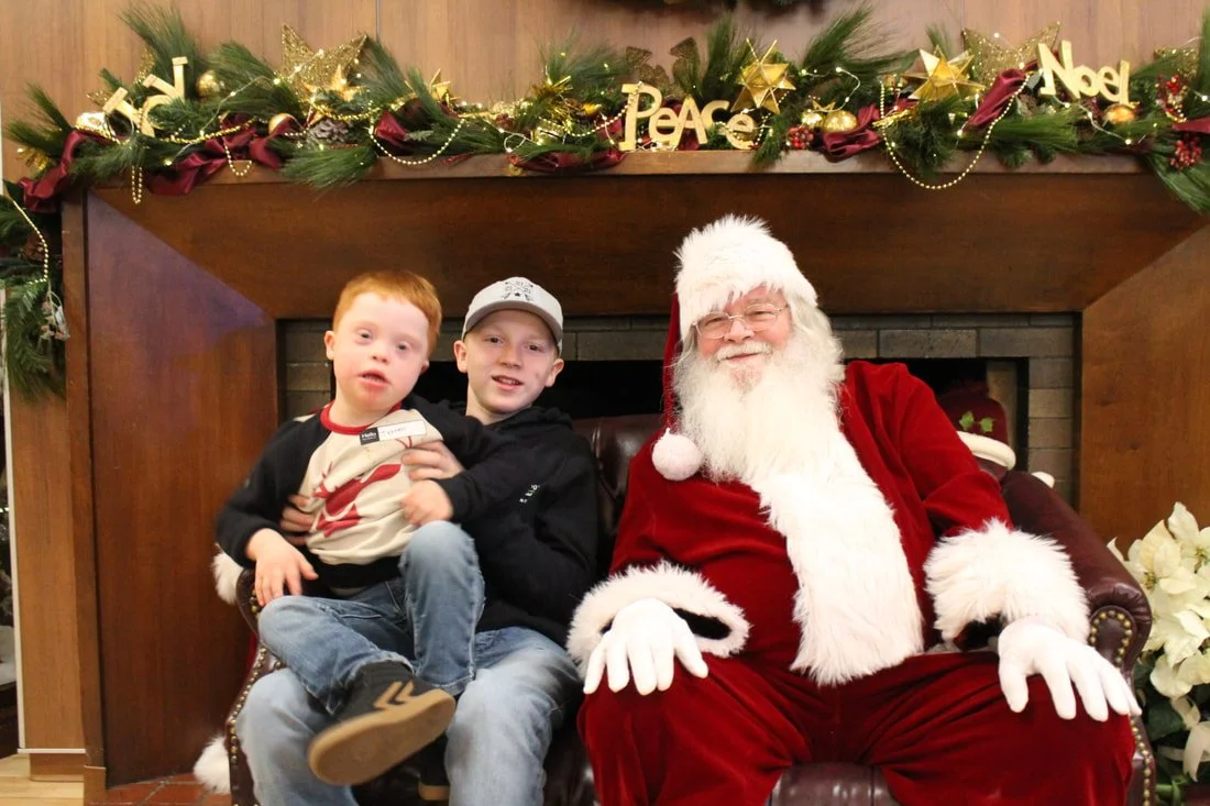 Two boys sitting next to Santa Claus on a decorated fireplace mantle with Christmas garland, gold ornaments, and signs that say 'Peace', 'Noel', and 'Merry'.