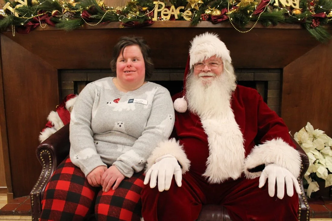 A woman sitting next to a man dressed as Santa Claus on a decorated Christmas backdrop, with festive ornaments and greenery overhead.