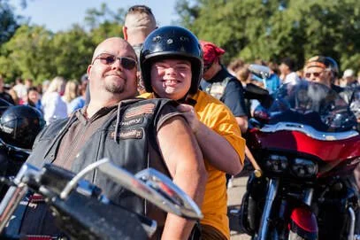 A man and a boy sitting on a motorcycle at a gathering with other motorcycles and people in the background, outdoors on a sunny day.