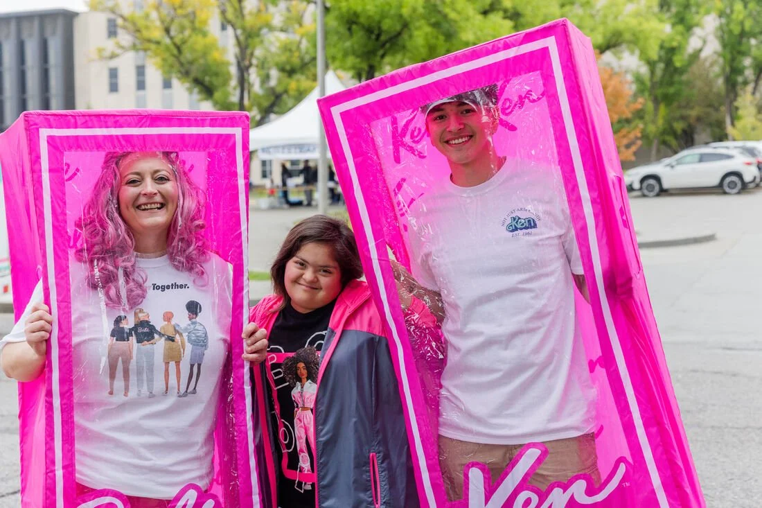 Three smiling people holding pink photo frames, outdoors in a parking lot, with trees and tents visible in the background.