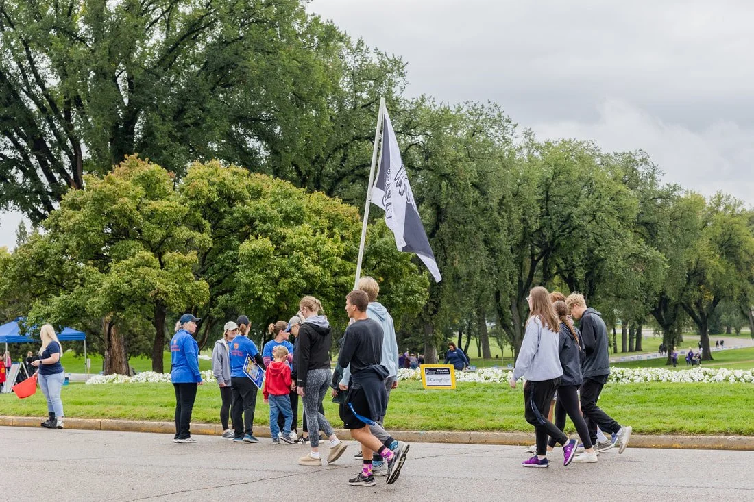 A group of people participating in a parade or march in a park on a cloudy day, with trees and greenery in the background.