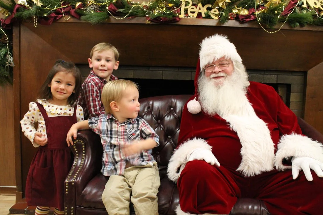 Three children sitting with Santa Claus on a leather sofa in front of a decorated fireplace with a Christmas garland and ornaments.