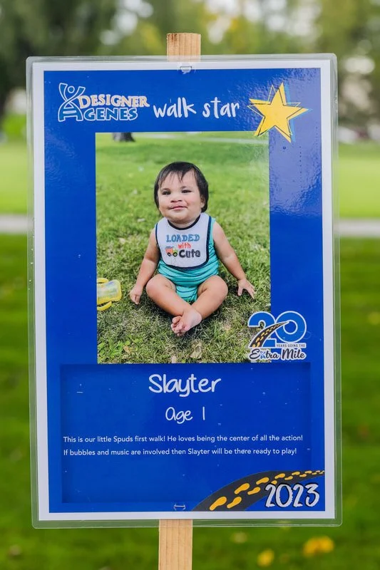 A young child sitting on grass inside a Blue Designer Genes walk star photo frame at a park. The frame has a yellow star and a celebration graphic with the number 20, indicating a milestone or anniversary. The child is wearing a white tank top with c