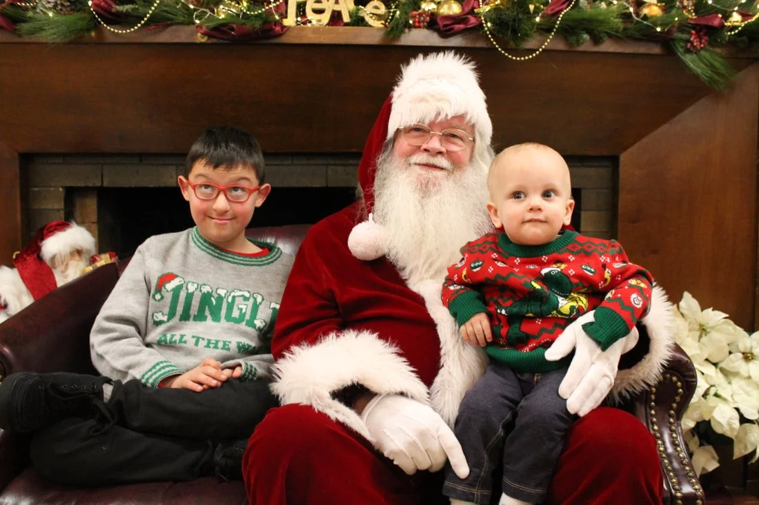 A man dressed as Santa Claus sitting on a bench with two children, one older and one younger, in front of a Christmas fireplace mantel decorated with greenery, gold beads, and ornaments.