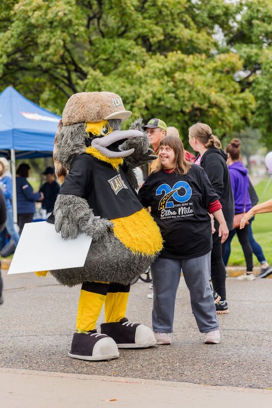 A person in a large bird mascot costume talking to a smiling woman at an outdoor event. The mascot is wearing a hat, shirt, and yellow accessories, and the woman is wearing a black T-shirt celebrating a 20th anniversary.