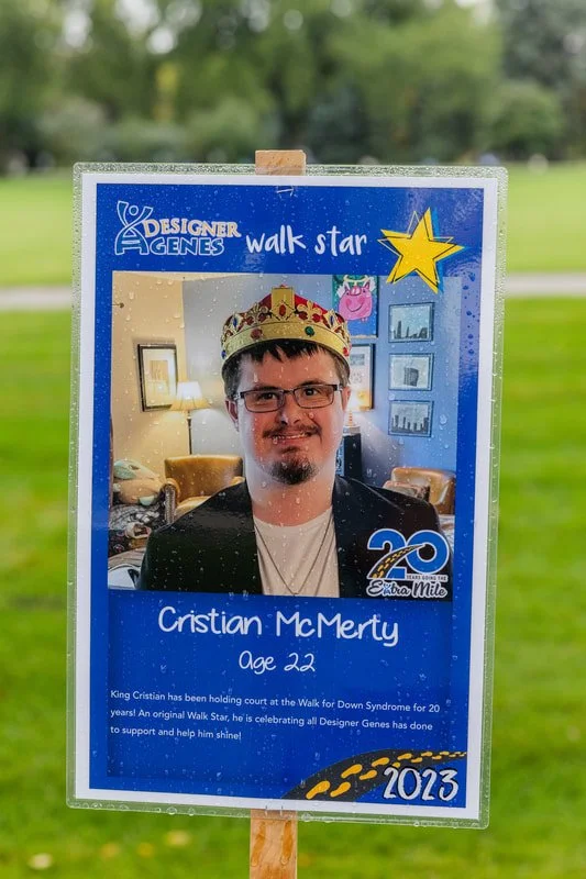 A sign celebrating Cristian McMerty's 22nd birthday and 20 years with Down Syndrome, featuring a photo of him wearing a crown, inside a living room with lamps, couches, and framed pictures, outdoors on grass.