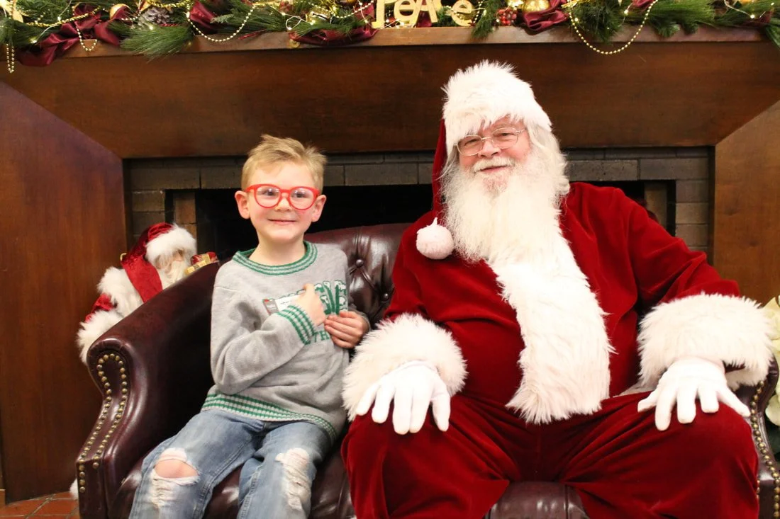 A young boy with glasses sitting next to Santa Claus on a leather sofa in a decorated Christmas setting.