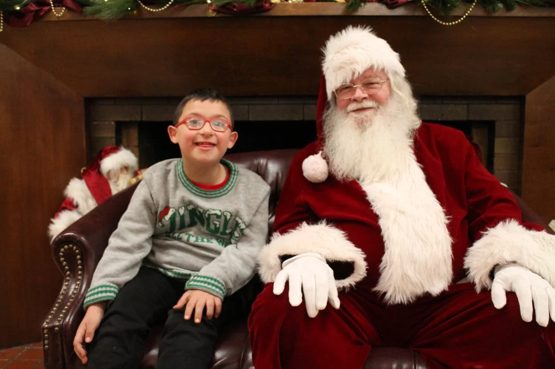 A boy with glasses and a gray sweater sitting beside a man dressed as Santa Claus, both smiling, in front of a fireplace decorated for Christmas.