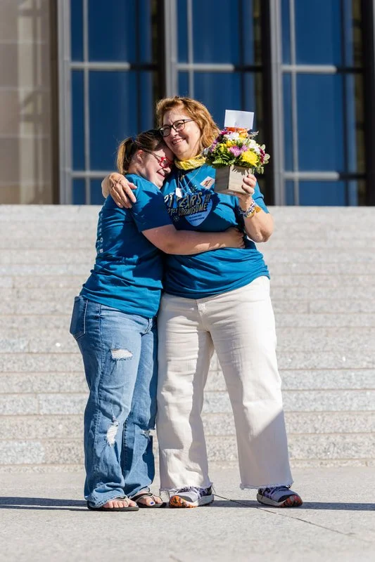 Two women hugging, one holding a bouquet of flowers, standing on steps outside a building with large glass windows.