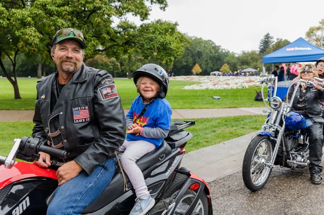 A man and a young girl sitting on a motorcycle in a park during daytime. The man is wearing a leather jacket, sunglasses, and has a beard. The girl is wearing a helmet and smiling. There are other motorcycles and people in the background, with trees 