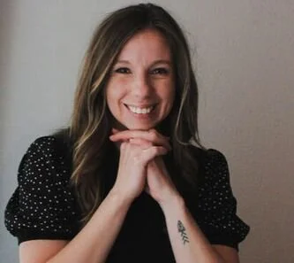 A woman with shoulder-length hair smiling, wearing a black polka dot top, with her hands near her chin, and a small tattoo on her wrist.