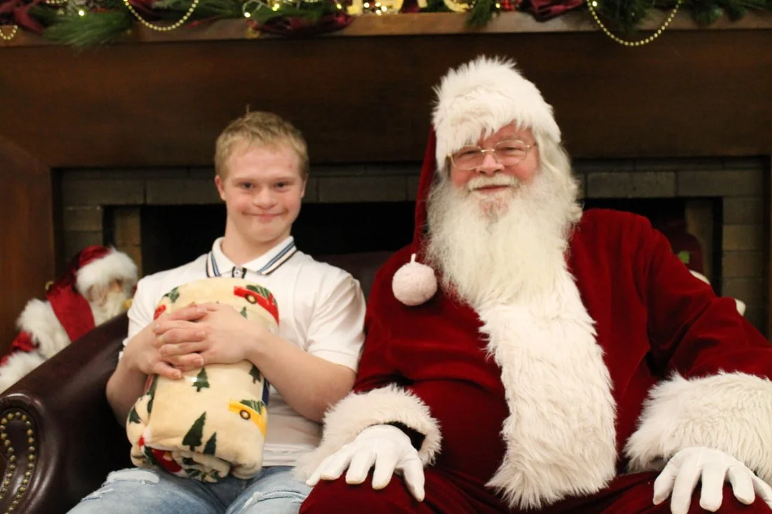 A young boy sitting next to Santa Claus, both smiling, during a Christmas celebration. The boy is holding a Christmas-themed gift, and Santa is dressed in his traditional red suit with white fur trim, in front of a decorated fireplace.