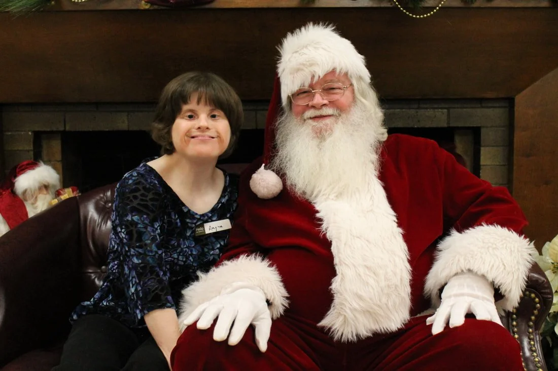 A young girl sitting next to Santa Claus, both smiling. Santa is dressed in a red velvet suit with white fur trim, wearing glasses and a Santa hat. They are in a room decorated for Christmas, with a fireplace and Christmas decorations visible in the 