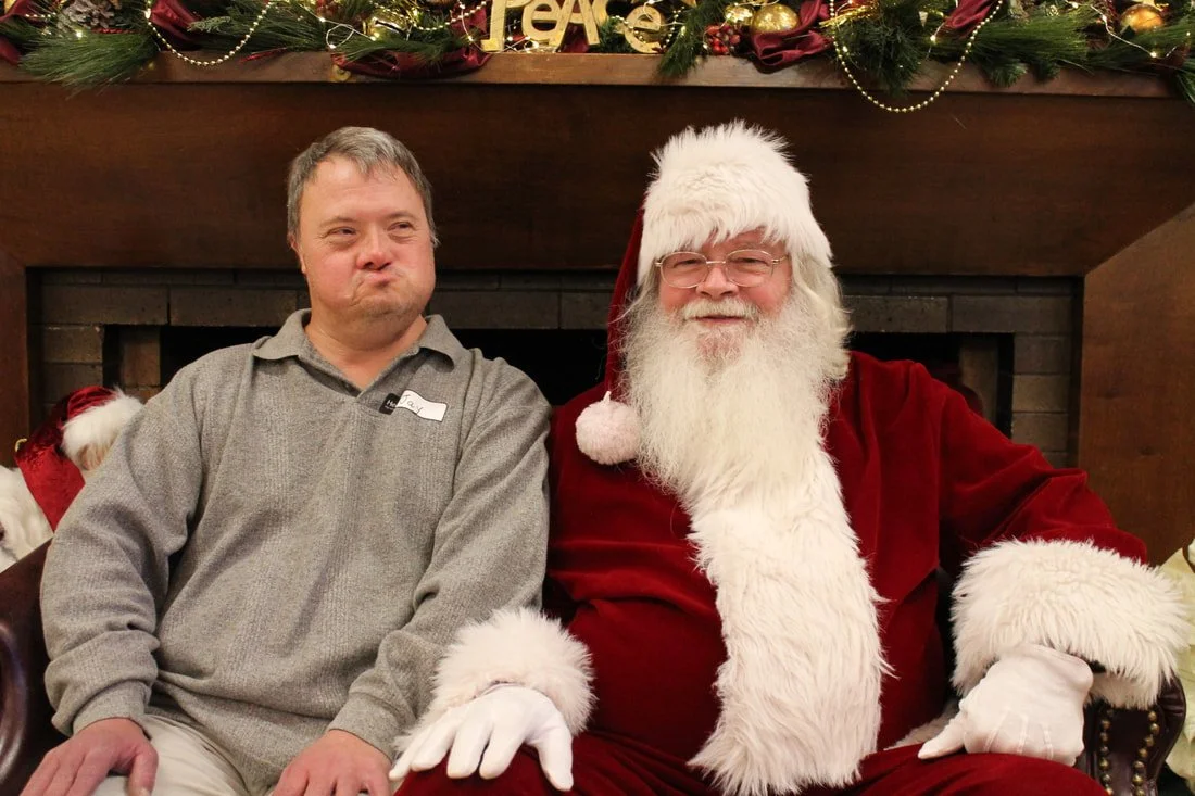 A man and Santa Claus sitting together in front of a fireplace decorated with Christmas garland and ornaments. The man is making a funny face, while Santa is smiling.