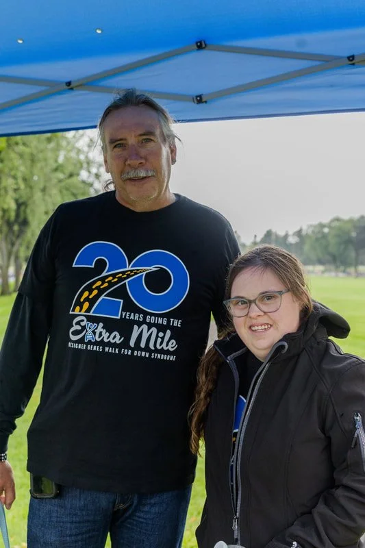 A middle-aged man with long hair and a beard, and a young woman with glasses, standing under a blue canopy in a park. The man is wearing a black T-shirt celebrating 20 years of walking for Down syndrome, and the young woman is wearing a black jacket.