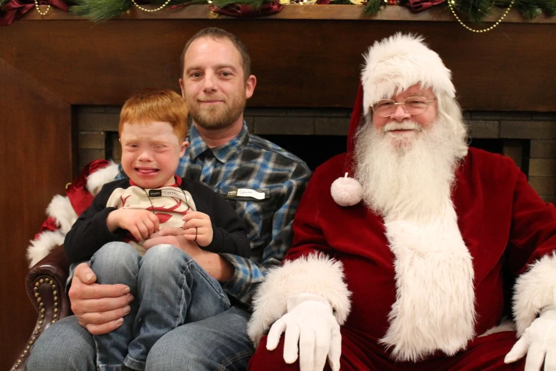 A young boy with red hair crying while sitting on a man's lap next to Santa Claus, who is dressed in a red suit with white trim, in front of a fireplace decorated for Christmas.