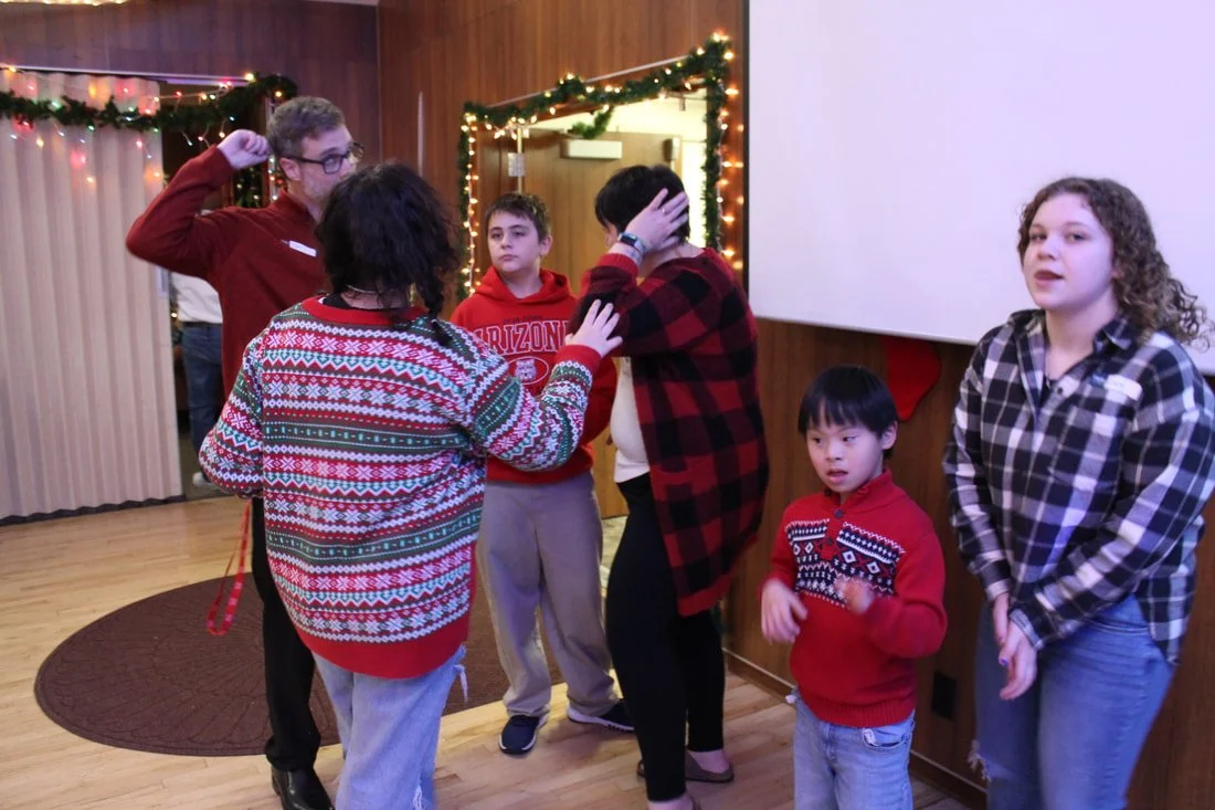 Group of children and adults gathered in a festive indoor setting with Christmas decorations and string lights.