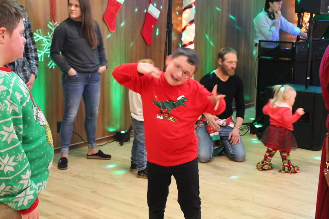 A group of people at a Christmas party, some dancing and others standing, with holiday decorations and stockings hanging on the wall.