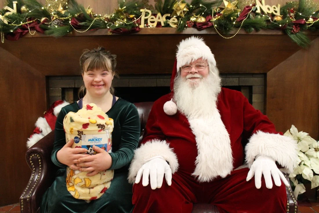 A young girl sitting on Santa Claus's lap holding a Christmas stocking, both smiling, in front of a decorated fireplace with holiday ornaments and greenery.