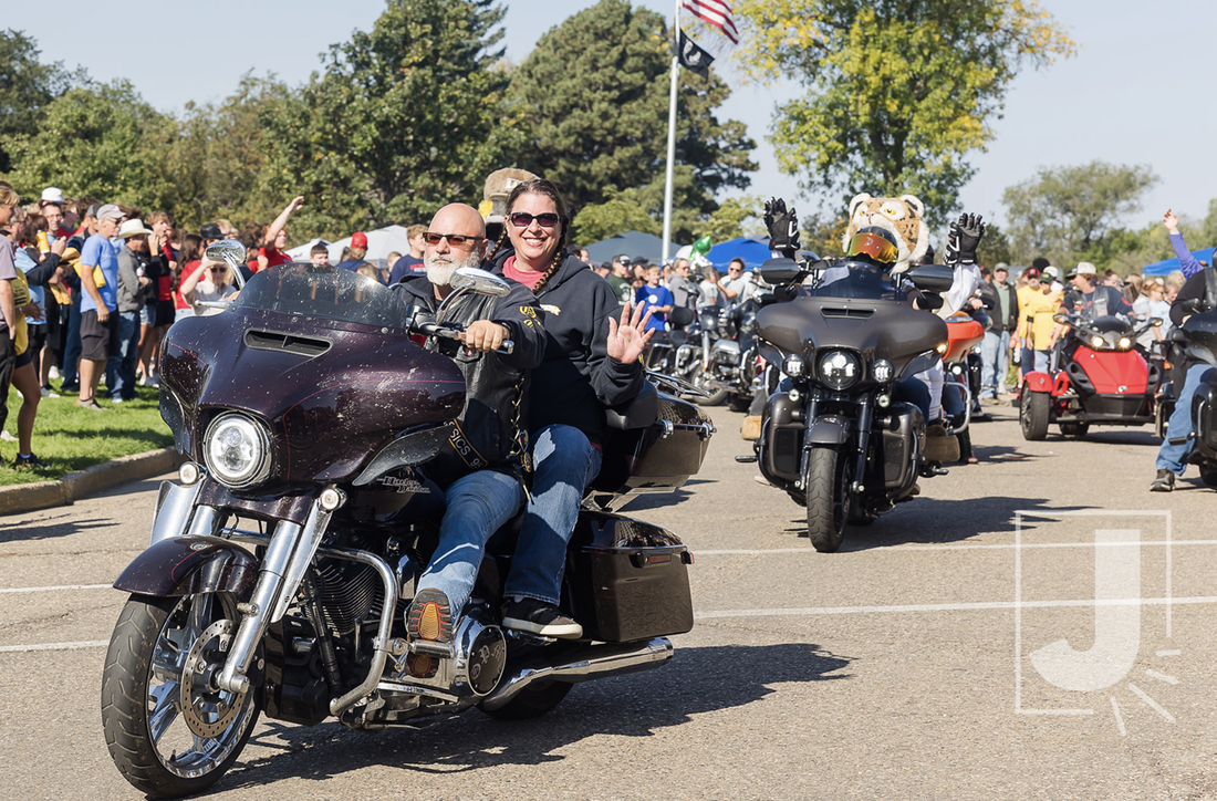 People riding and gathering around motorcycles and sidecars at an outdoor event, with a crowd in the background, and trees in the distance.