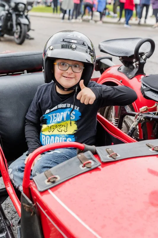 A young boy sitting in a red vintage car, wearing a black helmet and glasses, giving a thumbs up and smiling