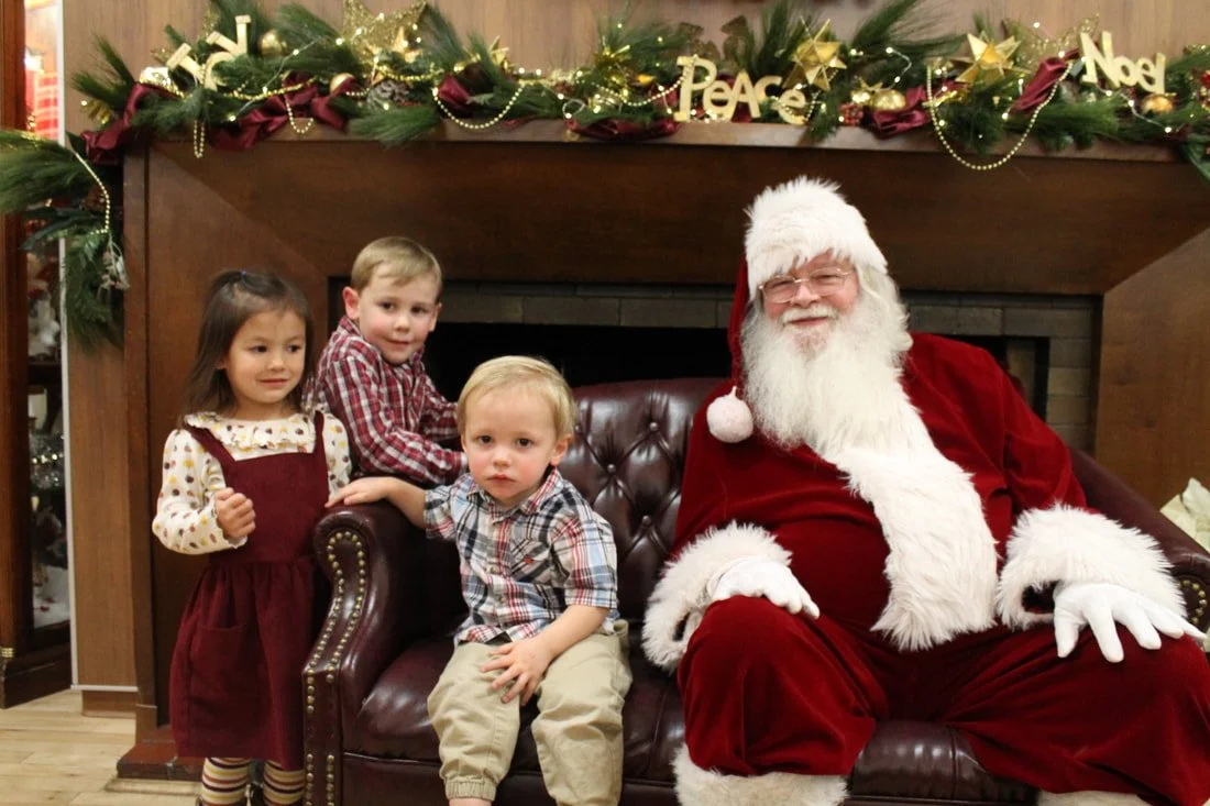 Four children sitting with Santa Claus on a Christmas-themed set with a decorated mantle in the background.