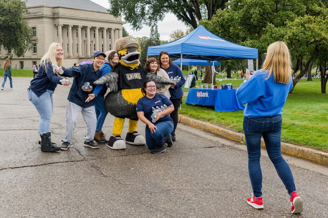 Group of people taking a photo with a mascot in front of a US Bank tent outdoors.