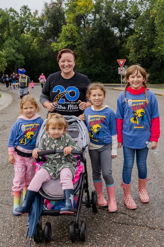 A group of five people, including four young girls and an adult woman, standing on a street during the day. The woman is smiling and wearing a black shirt with a 20th-anniversary logo. The girls are dressed casually and wearing colorful rain boots, a
