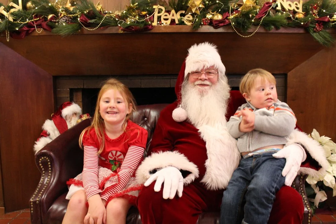 A young girl and a young boy sitting with Santa Claus on a fireplace hearth, decorated with Christmas ornaments, garland, and words like 'Peace' and 'Noel'.