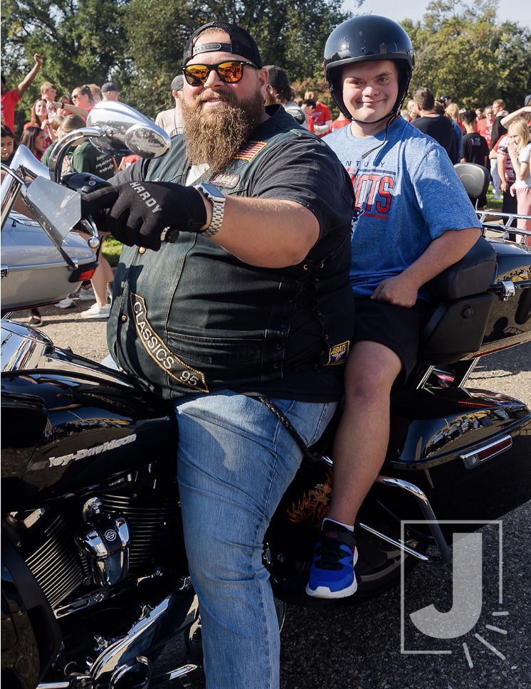 A man with a beard wearing sunglasses and a black vest sitting on a black Harley-Davidson motorcycle with a young boy sitting behind him, wearing a helmet and a blue T-shirt. They are at an outdoor event with a crowd of people in the background.