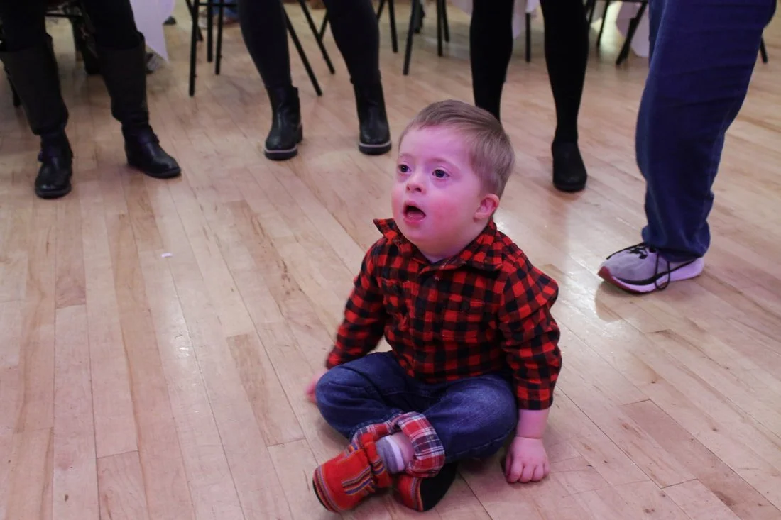A young boy sitting on a wooden floor with an expression of surprise or distress, wearing a red and black plaid shirt and jeans, surrounded by adults standing around.