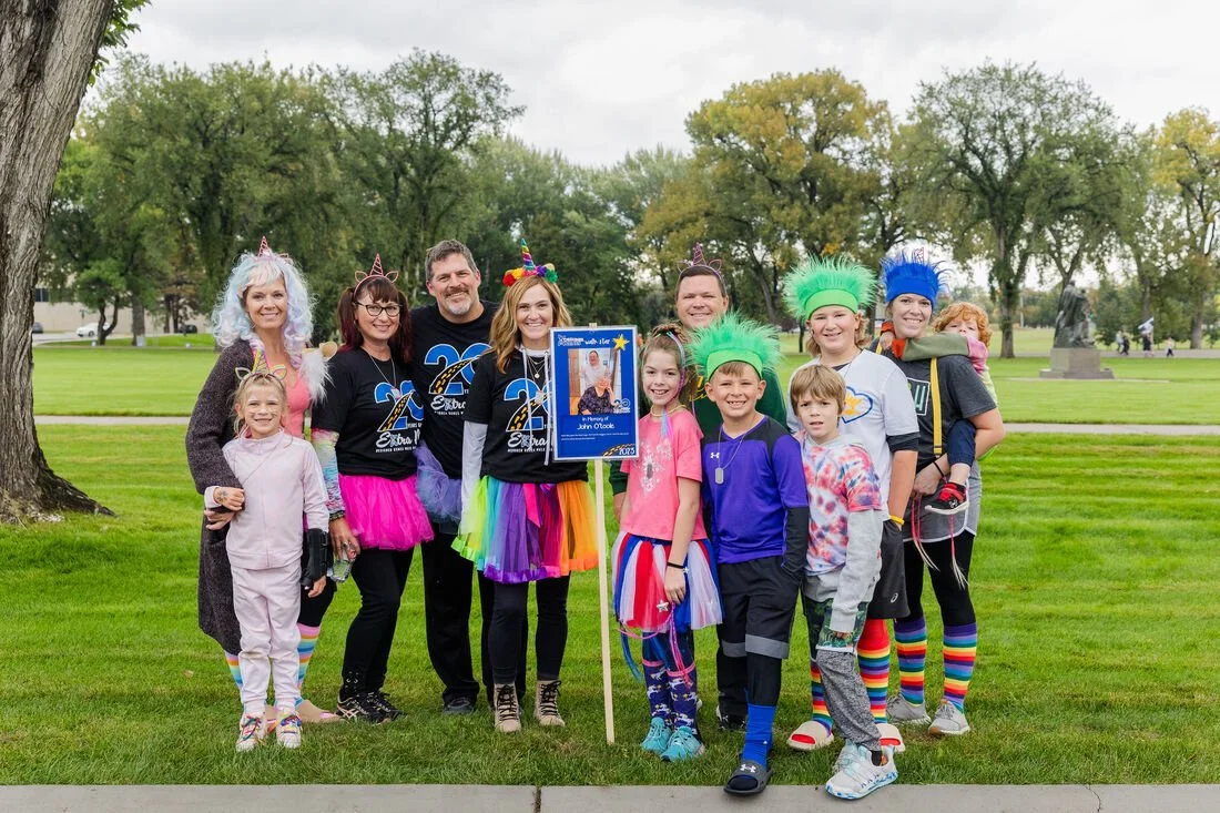 Group of children and adults dressed in colorful costumes and rainbow accessories, celebrating outdoors in a park, holding a framed photo and showing festive spirit.