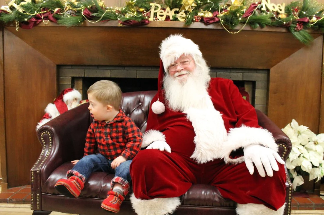 A young boy sitting on Santa Claus's lap on a brown leather couch in a festive Christmas setting.