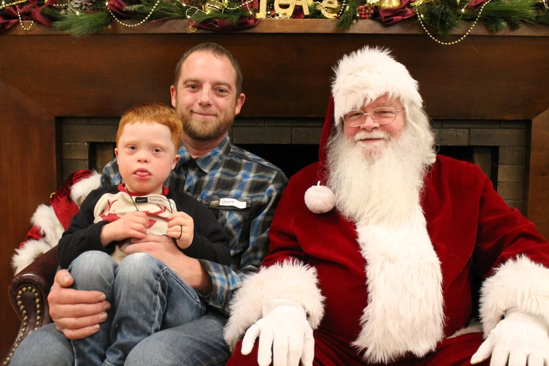 A young boy with red hair and a plaid shirt sitting on a man's lap, with a Santa Claus dressed in a red suit, white beard, and glasses, all sitting in front of a fireplace decorated for Christmas.
