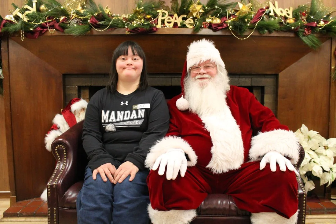 A girl sitting next to Santa Claus on a leather couch, inside a room decorated for Christmas with garlands, ornaments, and signs saying 'Peace' and 'Noel' on a wood-paneled wall above the fireplace.