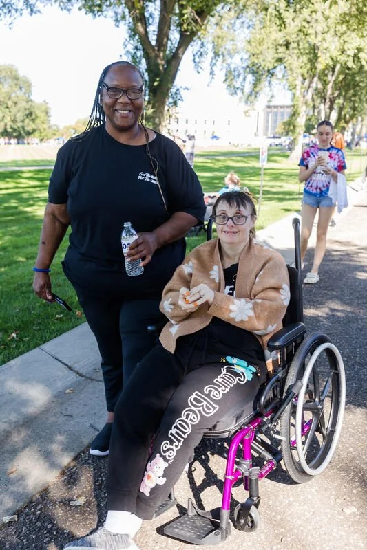 A woman with glasses and a brown blanket sitting in a wheelchair, smiling, and holding a snack, outdoors on a sunny day with trees in the background. Another woman with glasses and long hair stands beside her, holding a water bottle, also smiling, wi