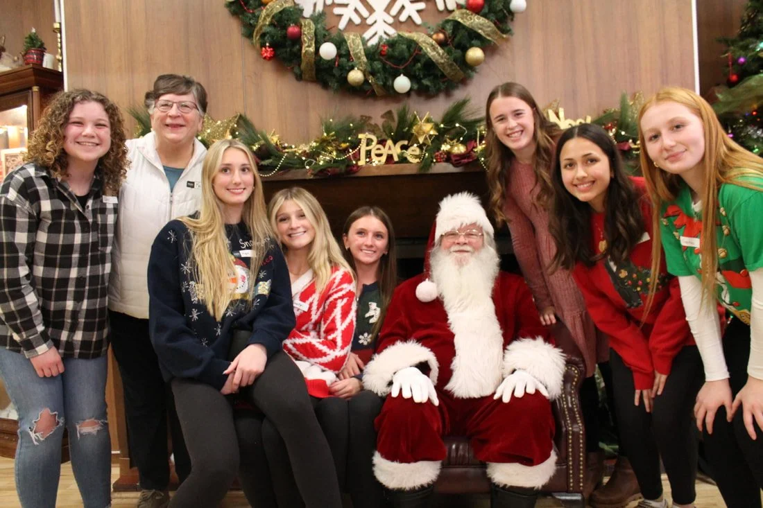 A group of eight young girls and one older woman gathered around a man dressed as Santa Claus in a festive indoor setting with Christmas decorations and a wreath on the wall.
