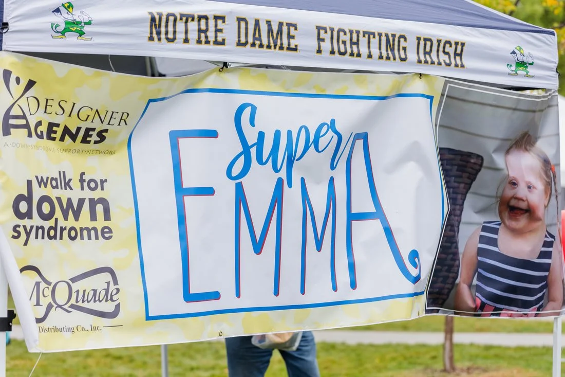 Banner at a charity walk event for Down syndrome awareness, featuring the words "Super Emma" and a picture of a smiling young girl with Down syndrome wearing a striped dress, under a tent with the text "Notre Dame Fighting Irish".