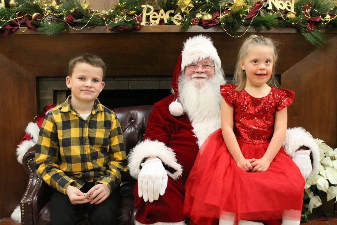 A boy and a girl sitting with Santa Claus in front of a Christmas fireplace mantel decorated with greenery, gold ornaments, and signs that spell 'Peace' and 'Noel'.