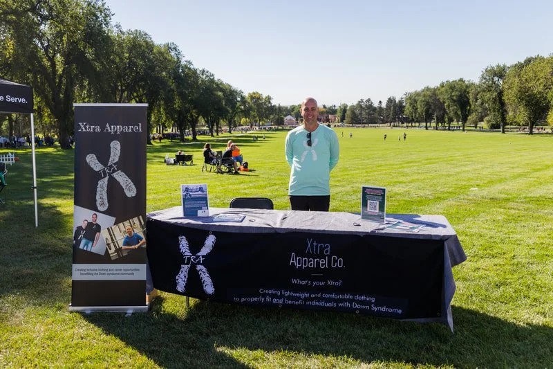 A man stands behind a table with a black tablecloth at an outdoor event in a park. The table has promotional materials for Xtra Apparel Co., which creates lightweight and comfortable clothing for individuals with Down Syndrome. A vertical banner with