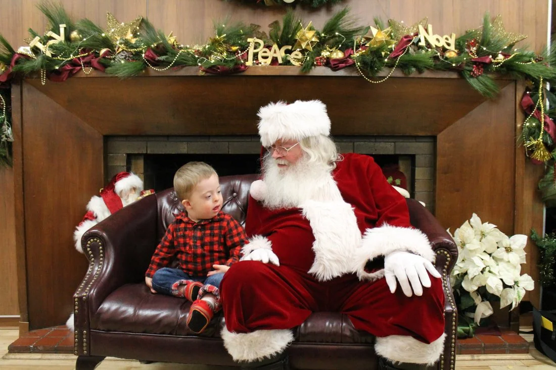 A young boy sitting on Santa Claus's lap during a Christmas celebration, decorated with festive garland, ornaments, and poinsettias.