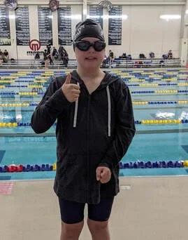 Young swimmer in swim goggles, cap, and jacket at an indoor swimming pool, giving a thumbs-up