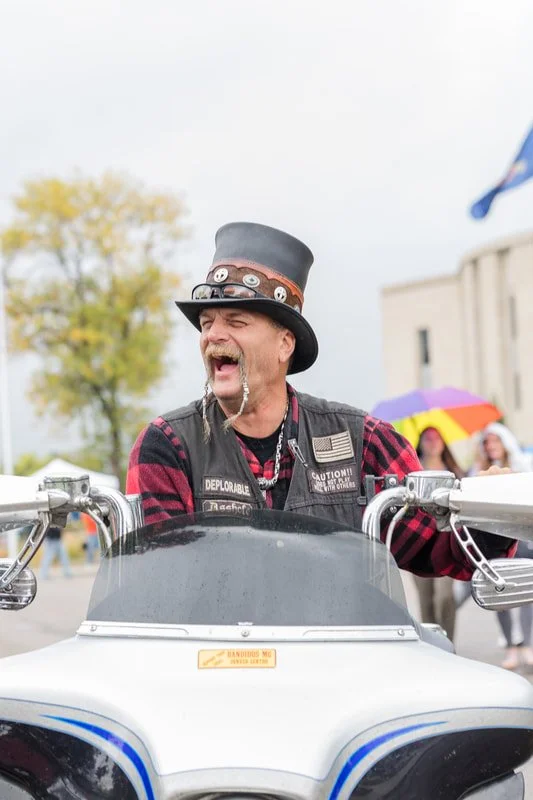 A man with a mustache, wearing a black hat with pins, a plaid red and black shirt, and a vest, laughing while sitting on a motorcycle.
