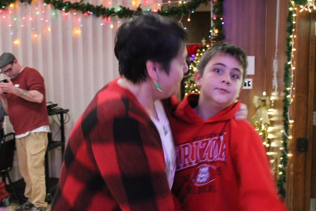 A woman and a boy sharing a hug at a Christmas celebration with holiday lights and decorations in the background.
