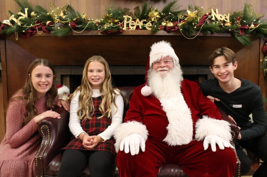Three children and a man dressed as Santa Claus sitting on a leather sofa in front of a fireplace decorated for Christmas.