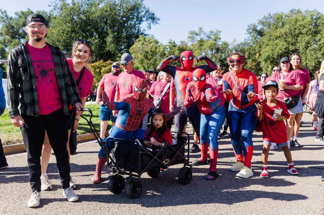 Group of people, including adults and children, gathered outdoors during daytime in Mankato, Minnesota, with some dressed as Spider-Man characters, and others wearing casual clothes. The scene appears festive and lively.